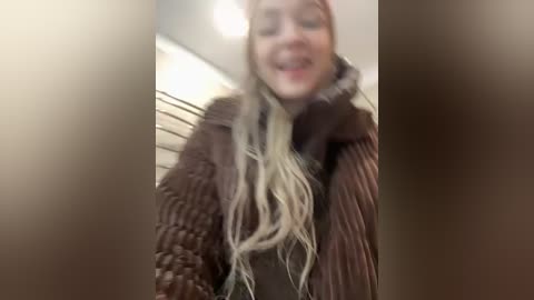 Media: A blurry video shows a smiling woman with long, wavy blonde hair, wearing a brown, textured sweater. She appears to be indoors, with a blurred background of vertical metal bars and beige walls.