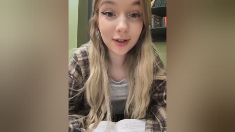 Media: Video of a young Caucasian woman with long blonde hair, wearing a plaid shirt, smiling while reading a book, indoors with a bookshelf in the background.