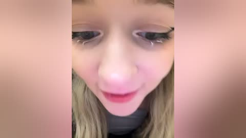 Media: Video of a young blonde girl with tear-streaked face, light skin, and slightly parted lips, viewed from a close-up angle, surrounded by blurred, pinkish background.