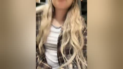 Media: A video of a young woman with long, wavy blonde hair wearing a plaid shirt over a white tank top, smiling slightly. The background is blurred, focusing on her upper body.