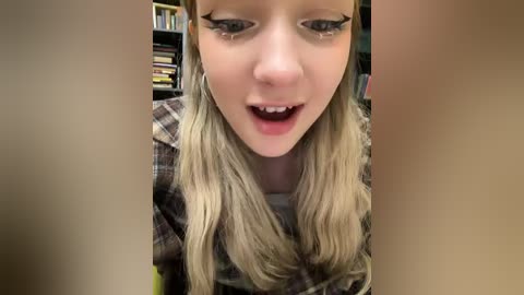 Media: Video of a young woman with long blonde hair, wearing black winged eyeliner, smiling with open mouth, sitting in a room with bookshelves and colorful books in the background.