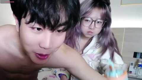 Media: Video of a young Asian man with short black hair, shirtless, leaning forward, and a young Asian woman with long purple hair, wearing glasses and a floral pajama top, in a bedroom.