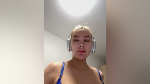 Media: A video of a light-skinned woman with platinum blonde hair, wearing silver headphones, sitting indoors with white walls and a ceiling light. She has a neutral expression, wearing a blue bra.