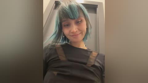 Media: A young woman with light skin and vibrant teal hair styled in a bob, wearing a black shirt with brown suspenders, videoed in a dimly lit hallway.