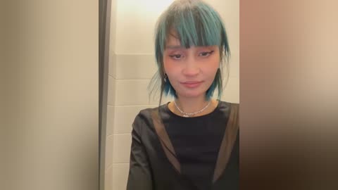 Media: Video of a young Asian woman with teal hair and bangs, wearing a black leather jacket, standing in a beige-tiled bathroom.