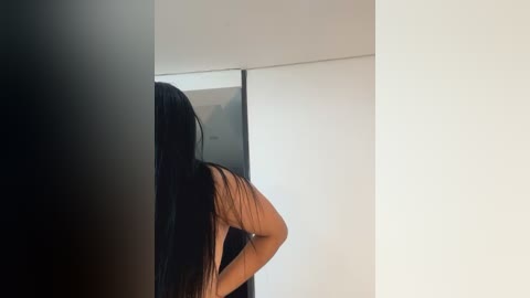 Media: A video of a woman with long, dark hair, wearing a brown top, standing in front of a mirrored wall, creating a reflective effect. The background is minimalist with soft, diffused light.