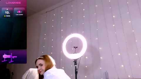 Media: Video of a young woman with fair skin and shoulder-length, light brown hair, smiling. She's wearing a white sweater, seated in front of a large, round, glowing ring light on a white backdrop with string lights.