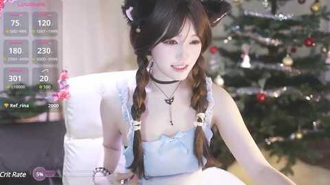 Media: Video of a young Asian woman with long brown hair in twin braids, wearing black cat ears, a choker, and a light blue dress, sitting in a cozy room with a decorated Christmas tree.