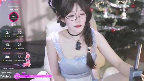 Media: Video of a fair-skinned Asian woman with black hair in braids, wearing glasses and a blue lace camisole, seated at a desk, with a blurred festive background.