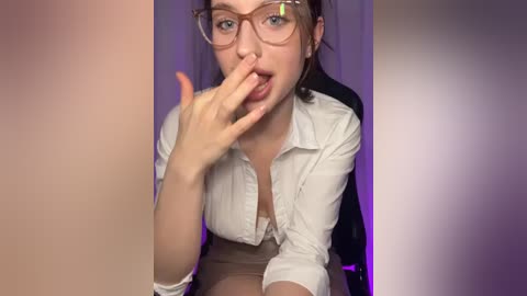 Media: Video of a young woman with fair skin, glasses, and brown hair, wearing a white blouse and beige skirt, playfully biting her finger against a purple background.