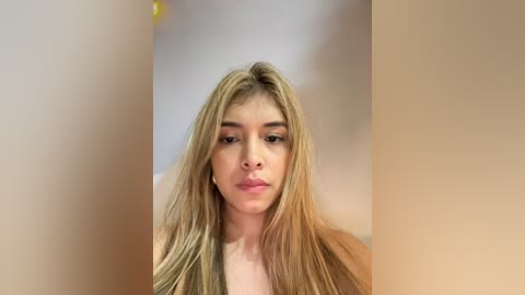 Media: A close-up video of a young woman with long, straight blonde hair, light skin, and a neutral expression. She's in focus, with blurred beige walls in the background.