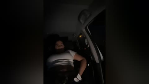 Media: A dimly-lit video of a woman with medium skin tone and dark hair, wearing a white crop top and black pants, leaning against the side of a car in a dimly lit parking lot.