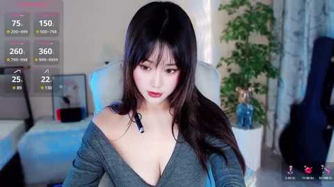 Media: Video of an Asian woman with long, dark hair and fair skin, wearing a grey off-shoulder top, sitting in a modern living room.