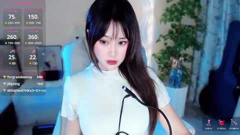 Media: Video of an Asian woman with long black hair, wearing a white turtleneck, sitting in front of a computer with a microphone. Background shows a green plant, blue curtains, and a guitar.