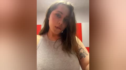 Media: Video of a young woman with fair skin, long brown hair, and a tattoo on her arm. She wears a light gray tank top and has a neutral expression. Background shows a red and white wall.