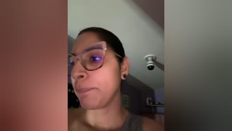 Media: A close-up video of a young woman with medium skin tone, short dark hair, and rectangular glasses. She wears a gray tank top and black earrings. The background features a dimly lit room with a ceiling light and blurred furniture.