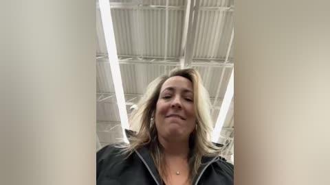 Media: Video of a smiling, light-skinned woman with shoulder-length blonde hair in a black hoodie, standing in an industrial, white ceiling-tiled room with fluorescent lighting.