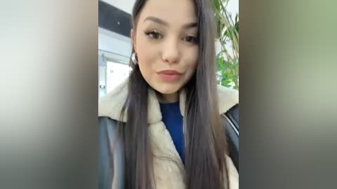 Media: A video of a young woman with long, straight, dark brown hair, wearing a blue top and a black leather jacket with a beige fur collar, indoors with green plants in the background.