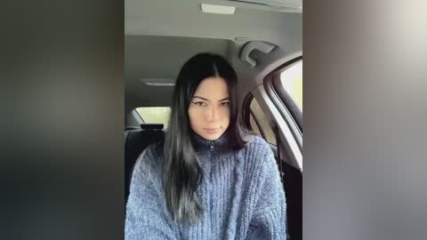 Media: Video of a young Asian woman with long, straight black hair, wearing a gray, knitted sweater, sitting in the backseat of a car with gray interior.