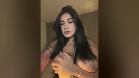 Media: A video of a topless woman with long black hair, fair skin, and tattoos, covering her breasts with her hands. She has a confident expression, wearing bold red lipstick.