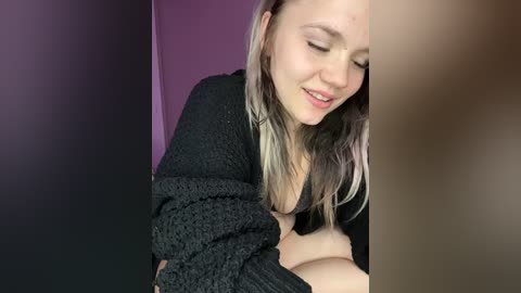 Media: Video of a smiling, fair-skinned woman with light blonde hair, wearing a black, textured sweater, sitting on a purple wall, with blurred background.