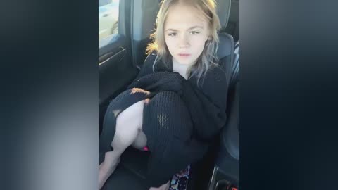 Media: A video of a young woman with blonde hair, wearing a black knit sweater, sitting in a car. She has fair skin and is leaning forward, partially exposed in a revealing pose.