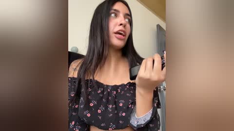 Media: Video of a young woman with long black hair, wearing a floral off-shoulder top, applying makeup in front of a mirror.