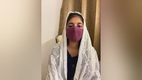 Media: Video of a young woman with medium skin tone and dark hair wearing a maroon mask, white headscarf, and navy blouse, standing in a room with beige walls and gold curtains.