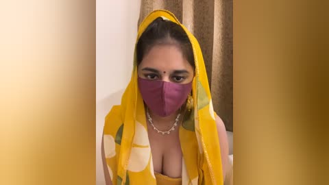 Media: Video of a South Asian woman in a yellow saree with a purple face mask, her dark hair tied back, indoors with beige curtains.