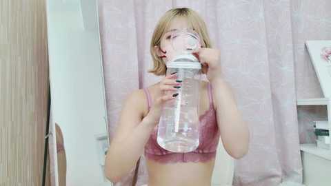 Media: A video of a young woman with short blonde hair, wearing a pink lace bra, drinking from a clear plastic water bottle. She is indoors, in a room with light-colored walls, a white cabinet, and a pink floral curtain.