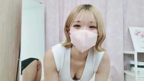 Media: A video of a young, slender East Asian woman with shoulder-length blonde hair, wearing a white top and a pink face mask. She has a shy expression. Background features a white shelf and a light pink curtain.