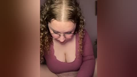 Media: Video of a Caucasian woman with curly brown hair, wearing glasses, and a low-cut maroon top, bending over to look at a phone, partially obscured by two vertical brown objects.