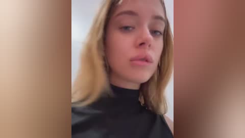 Media: A close-up video of a young woman with shoulder-length blonde hair, wearing a black turtleneck. Her facial expression is neutral, and the image is blurred with a reddish tint on the right side.