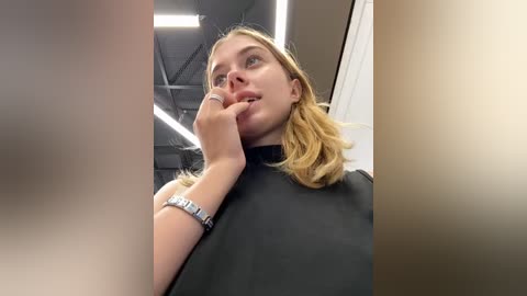 Media: Video of a young Caucasian woman with shoulder-length blonde hair, wearing a black robe, standing indoors with a thoughtful expression, holding her chin, and wearing a silver bracelet.