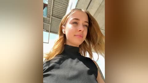 Media: Video of a young Caucasian woman with long, light brown hair, wearing a black turtleneck sweater. She stands against a modern building with large windows and a textured ceiling, her expression calm and composed.