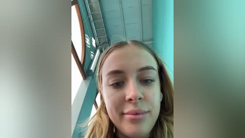 Media: Video of a young Caucasian woman with fair skin and blonde hair, wearing a white top, standing in a modern indoor space with light blue walls and metal railings.