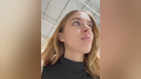 Media: Video of a young, fair-skinned woman with long, wavy, light brown hair, wearing a black top, looking upward, set against a white, grid-patterned ceiling.