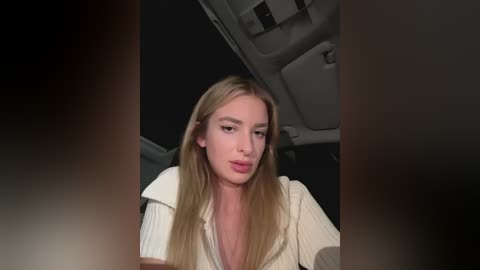 Media: Video of a young woman with long, straight blonde hair, fair skin, and light makeup, wearing a cream-colored sweater. She's seated in a car with dark interior.