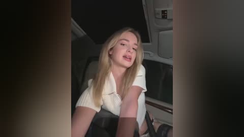 Media: Video of a young blonde woman with long hair, wearing a white polo shirt, leaning against a car seat, dimly lit interior, slightly blurred.