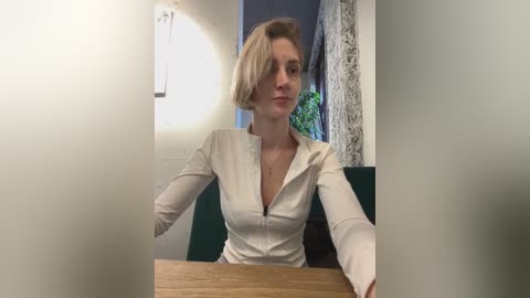 Media: Video of a blonde woman with a short bob haircut, wearing a white, long-sleeved, zip-up blouse, seated at a wooden table in a modern, well-lit room with green and white decor.