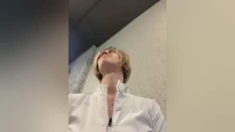 Media: Video of a young man with short, light brown hair, wearing a white shirt, standing in a dimly lit, beige-walled corridor, looking upward with a contemplative expression.