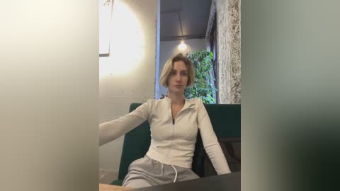 Media: Video of a blonde Caucasian woman with a slender build, wearing a beige zip-up sweater and grey sweatpants, seated in a modern, green upholstered chair in a minimalist, warmly lit room.