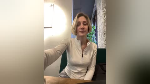 Media: Video of a blonde woman with a fair complexion, wearing a white long-sleeved shirt, sitting in a green upholstered chair in a dimly lit room with a wall-mounted light and a partially visible window.