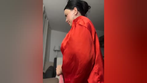 Media: Video of a woman with dark hair in a bun, wearing a red satin robe, standing in a dimly lit room with a gray wall and a partially visible bed.