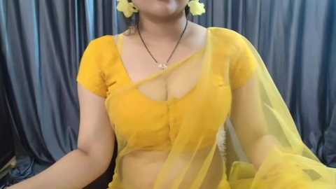 Media: Video of a South Asian woman with medium skin tone wearing a vibrant yellow saree, adorned with yellow flowers and a matching blouse, seated against a gray curtain backdrop.