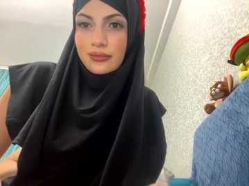 Media: Video of a young woman in a black hijab with a red flower, smiling, in a room with a blue towel, beige wallpaper, and stuffed animals.