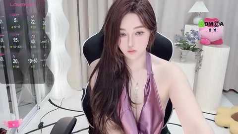 Media: A video of an Asian woman with long brown hair, wearing a purple halter top, seated in a black office chair in a modern room with white walls, curtains, and a calendar.