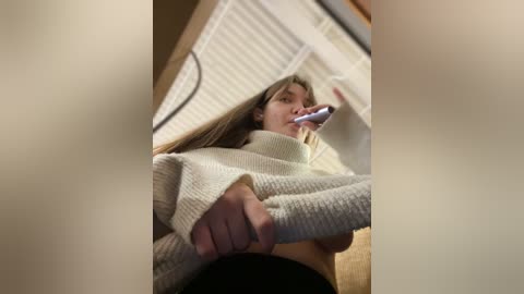 Media: Video of a young woman with long blonde hair brushing her teeth, wearing a cream-colored sweater. She is indoors with white blinds in the background.