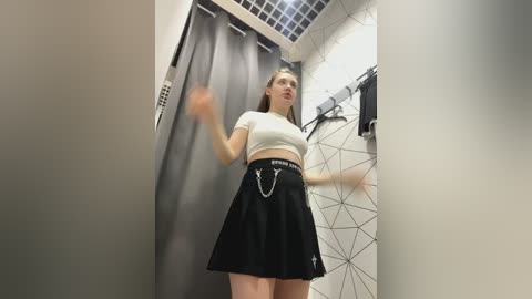 Media: Video of a young Caucasian woman with straight blonde hair, wearing a white crop top and black pleated skirt with chain belt, standing in a modern, geometric-patterned tiled dressing room.