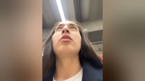 Media: Video of a woman with long, straight brown hair, wearing a dark blazer, looking upward with a neutral expression in a modern indoor setting with a gray ceiling and fluorescent lights.